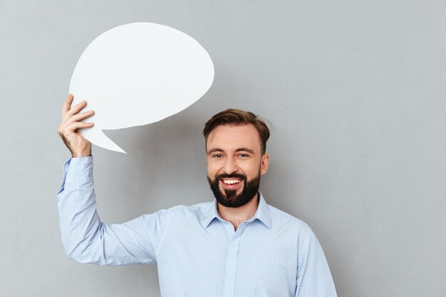 Heureux homme barbu dans des vêtements d'affaires tenant une bulle de dialogue vide