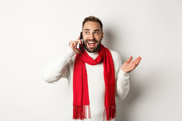 Heureux homme barbu souhaitant joyeux noël au téléphone, appelant quelqu'un et parlant, debout en pull avec écharpe rouge, fond blanc.