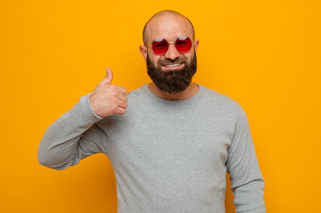Heureux homme barbu en sweat-shirt gris portant des lunettes rouges à sourire montrant le pouce vers le haut