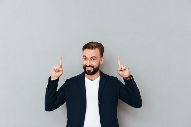 Heureux homme barbu en vêtements busines pointant vers le haut et regardant la caméra sur gris