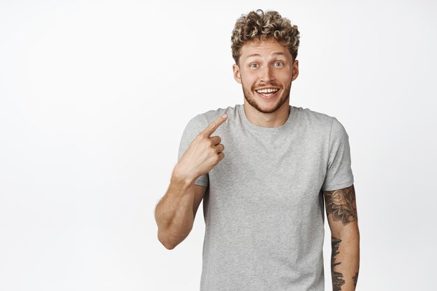 Heureux homme blond pointant le doigt vers lui-même avec un sourire excité étant choisi pour gagner un prix debout en t-shirt gris sur fond blanc