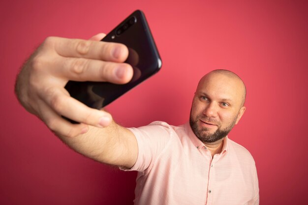 Heureux homme chauve d'âge moyen portant un t-shirt rose prendre un selfie isolé sur rose