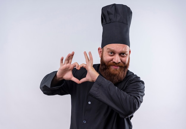 Photo gratuite un heureux homme chef barbu en uniforme noir montrant signe de forme de coeur avec les mains sur un mur blanc