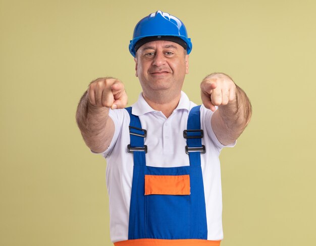 Heureux homme constructeur adulte en points uniformes à l'avant avec deux mains isolé sur mur vert olive