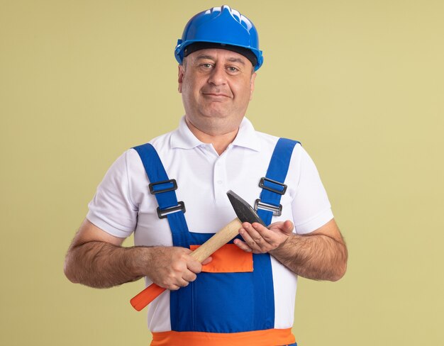 Heureux homme constructeur adulte en uniforme détient marteau isolé sur mur vert olive