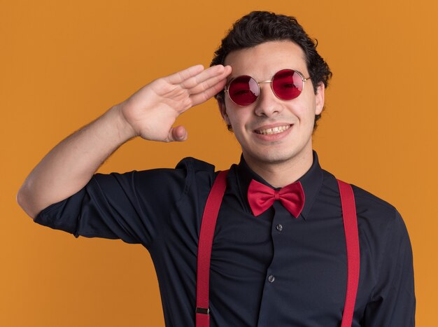 Heureux homme élégant avec noeud papillon portant des lunettes et des bretelles à l'avant avec le sourire sur le visage saluant debout sur le mur orange