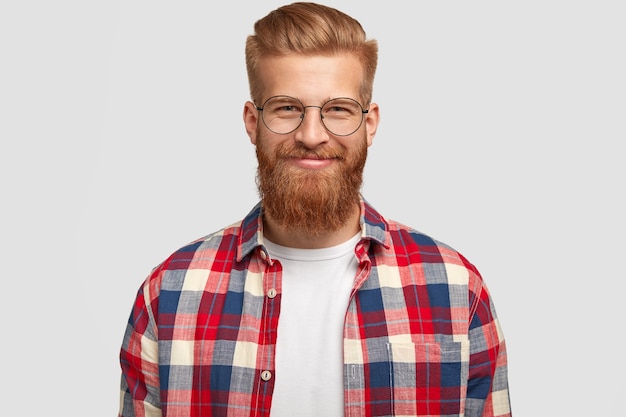 Heureux homme gingembre avec une expression heureuse, porte des lunettes et une chemise à carreaux à la mode, se réjouit du projet réussi, pose seul contre un mur blanc. Les gens, les émotions, le style de vie