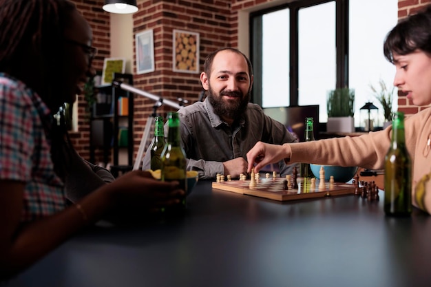 Heureux homme jouant aux échecs avec des amis multiethniques à la maison dans le salon. Diverses personnes assises à table et s'amusant en jouant ensemble à des jeux de société et en dégustant des collations et des boissons.