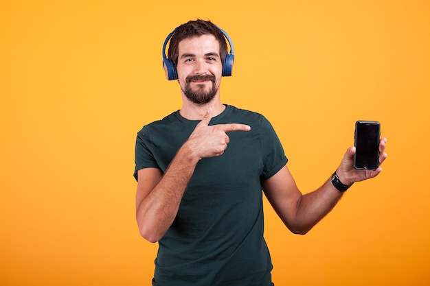 Heureux homme joyeux portant des écouteurs et pointant sur son smartphone isolé sur fond orange en studio.