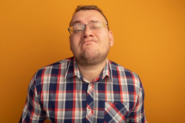 Heureux homme à lunettes et chemise à carreaux avec une expression confiante debout sur un mur orange