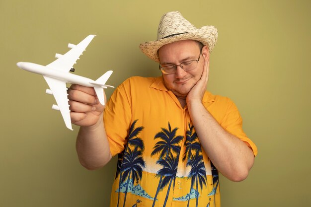 Heureux homme à lunettes portant une chemise orange en chapeau d'été tenant un avion jouet souriant avec la main sur sa joue debout sur le mur vert