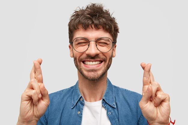 Heureux homme mal rasé avec un large sourire, montre des dents blanches, croise les doigts pour la bonne chance, être dans un esprit élevé se tient sur un mur blanc porte une chemise en jean à la mode. Un mec positif fait un geste d'espoir