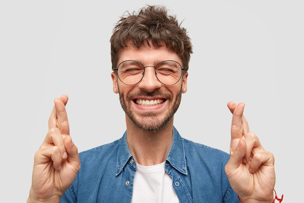 Heureux homme mal rasé avec un large sourire, montre des dents blanches, croise les doigts pour la bonne chance, être dans un esprit élevé se tient sur un mur blanc porte une chemise en jean à la mode. Un mec positif fait un geste d'espoir