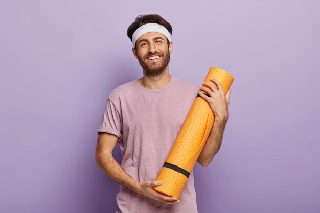 Heureux homme mal rasé tient un tapis de fitness roulé, satisfait après le cours de yoga