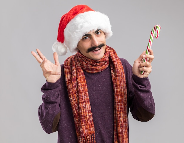 Heureux homme moustachu portant un bonnet de noel avec une écharpe chaude autour du cou tenant une canne en bonbon souriant joyeusement debout sur un mur blanc