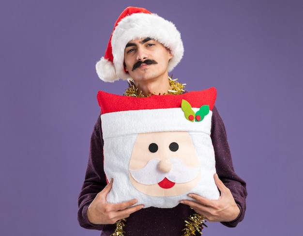 Heureux Homme Moustachu Portant Un Bonnet De Noel De Noël Avec Des Guirlandes Autour Du Cou Tenant Un Oreiller De Noël Avec Un Sourire Sur Le Visage Debout Sur Un Mur Violet