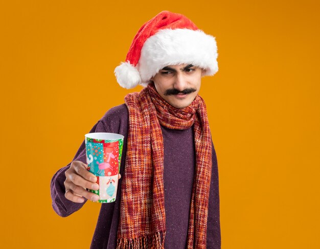 Heureux homme moustachu portant chapeau de père Noël avec écharpe chaude autour de son cou tenant une tasse de papier coloré en regardant avec le sourire sur le visage debout sur fond orange