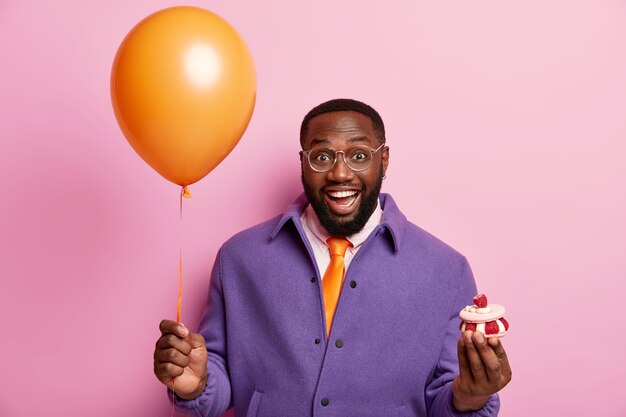 Heureux homme noir souriant tient ballon à air et petit petit gâteau, va féliciter un collègue avec anniversaire, a la bonne humeur