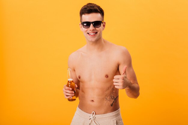 Heureux homme nu en short et lunettes de soleil tenant une bouteille de bière et montrant le pouce vers le haut tout en regardant la caméra sur jaune