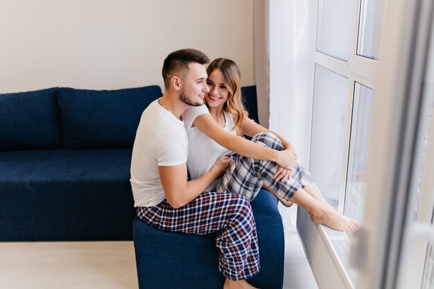 Heureux homme en pantalon à carreaux regardant la fenêtre. Charmante femme blonde au repos à la maison en bonjour.