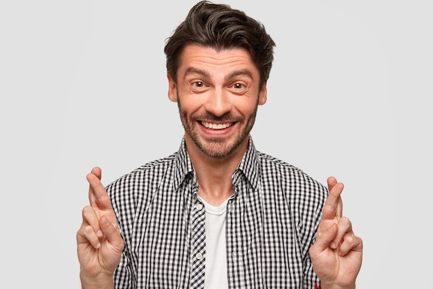 Heureux homme de race blanche avec coupe de cheveux à la mode, gestes à l'intérieur, soulève les doigts croisés, croit en la bonne chance, sourit sincèrement, porte une chemise à carreaux à la mode, isolée sur un mur blanc. Attente