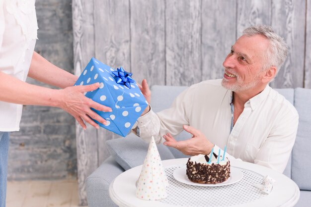 Heureux homme recevant un cadeau d&#39;anniversaire de sa femme près de gâteau et chapeau de fête sur table