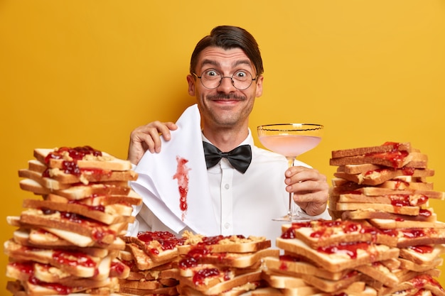 Heureux homme ringard élégant boit un cocktail, vêtu d'une chemise blanche avec noeud papillon, étant en banquet, tient une serviette blanche comme neige sale avec de la confiture, pose près de tas de pain, isolé sur mur jaune