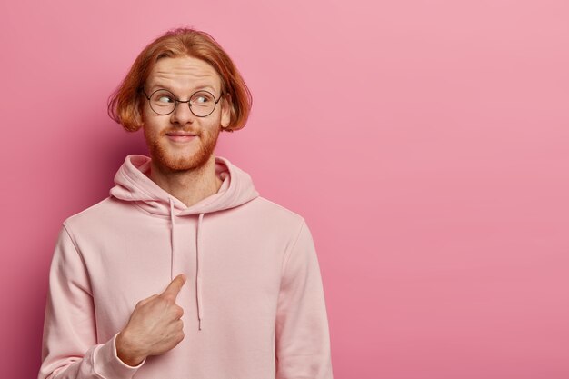 Heureux homme rousse avec une coiffure bob et des cheveux roux, se montre du doigt et regarde avec émerveillement de côté, porte des lunettes optiques et un sweat-shirt, demande si tu veux dire moi, pose contre un mur pastel rose