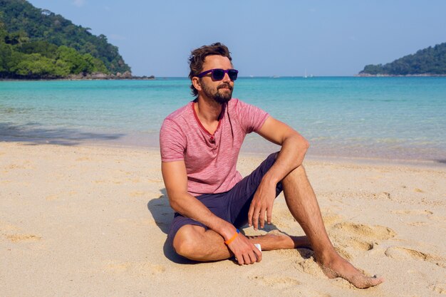 Heureux homme séduisant dans des lunettes de soleil élégantes avec barbe à l'aide de téléphone portable à la plage