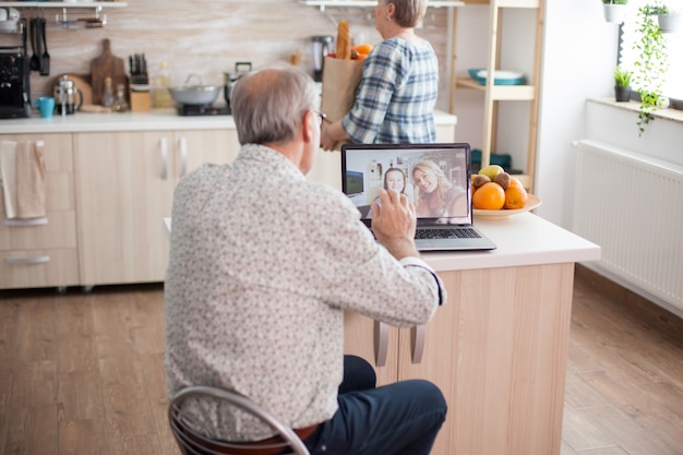 Heureux homme senior saluant sa nièce lors d'une vidéoconférence avec sa famille utilisant un ordinateur portable dans la cuisine. Appel en ligne avec sa fille. Personne âgée utilisant la technologie Web Internet en ligne de communication moderne.