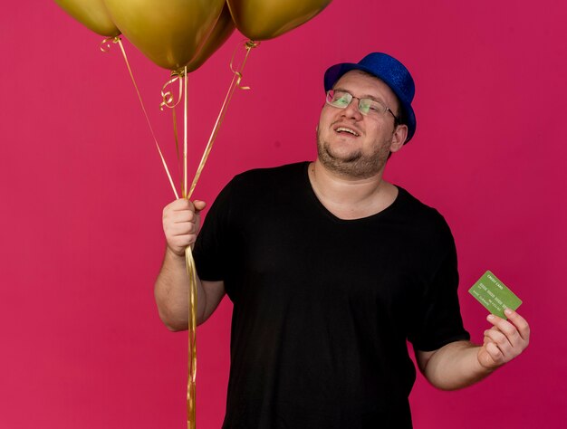 Photo gratuite heureux homme slave adulte dans des lunettes optiques portant un chapeau de fête bleu détient des ballons à l'hélium et une carte de crédit