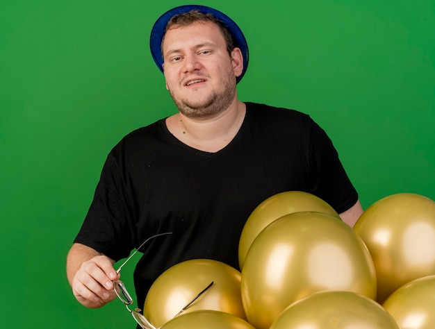 Heureux homme slave adulte détient des lunettes optiques portant un chapeau de fête bleu debout avec des ballons à l'hélium