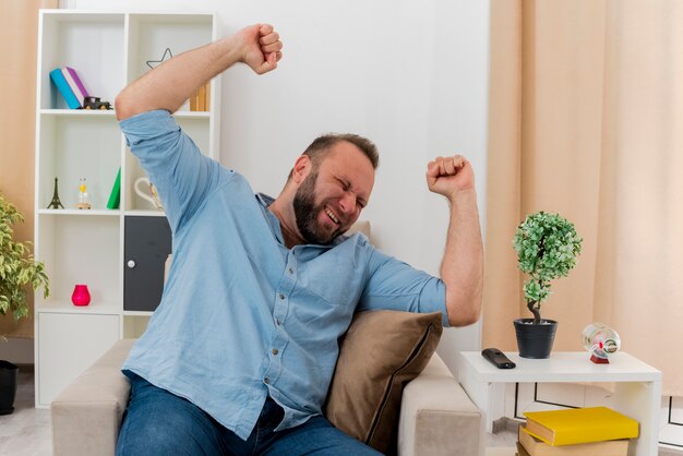 Heureux homme slave adulte est assis sur un fauteuil en levant les poings à l'intérieur du salon