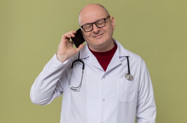 Heureux homme slave adulte avec des lunettes optiques en uniforme de médecin avec stéthoscope parlant au téléphone