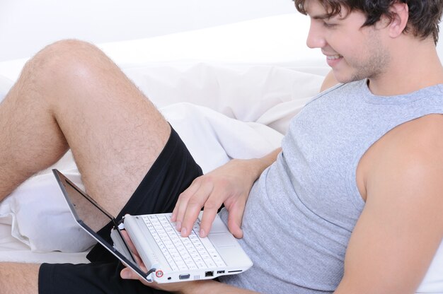 Heureux homme souriant en tapant sur un ordinateur portable au lit - vue de profil
