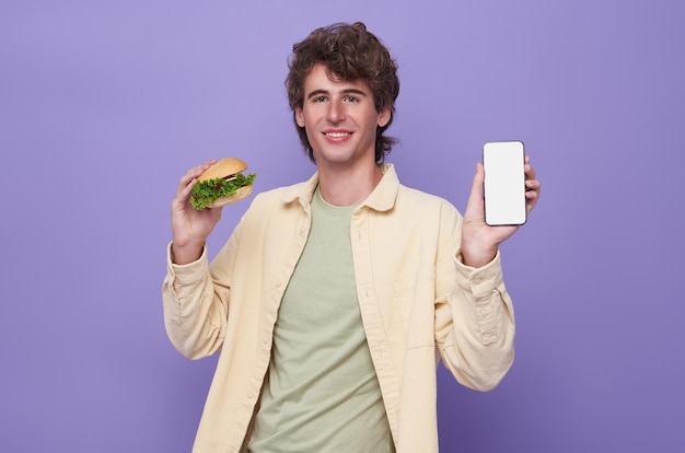 Heureux homme tenant un hamburger et montrant l'application de livraison de nourriture sur téléphone mobile pour commander le déjeuner