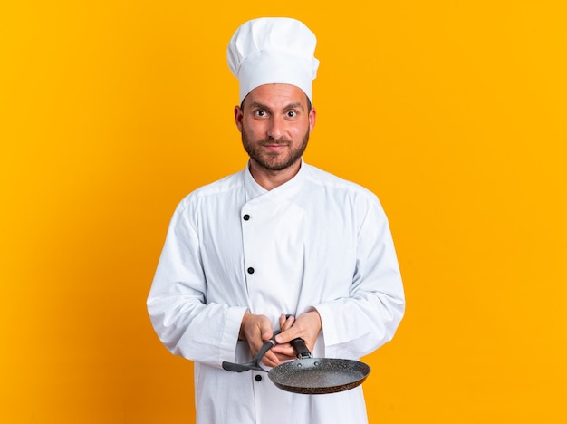 Heureux et impressionné jeune cuisinier caucasien en uniforme de chef et casquette tenant une spatule et une poêle à frire regardant la caméra isolée sur un mur orange avec espace de copie