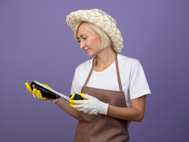 Heureux Jardinier Blonde D'âge Moyen Femme En Uniforme Portant Un Chapeau Et Des Gants De Jardinage Regardant Et Mesurant L'aubergine Avec Un Mètre Ruban