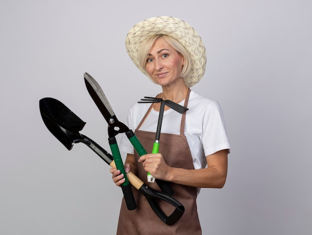 Heureux jardinier blonde d'âge moyen en uniforme portant un chapeau regardant à l'avant tenant des outils de jardinier isolés sur un mur blanc avec espace pour copie
