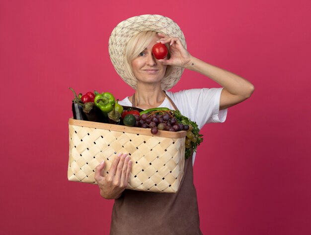 Heureux jardinier blonde d'âge moyen en uniforme portant un chapeau tenant un panier de légumes et de tomates devant les yeux regardant à l'avant isolé sur un mur cramoisi