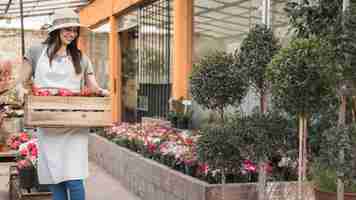 Photo gratuite heureux jardinier femelle transportant une caisse en bois pleine de fleurs rouges en serre