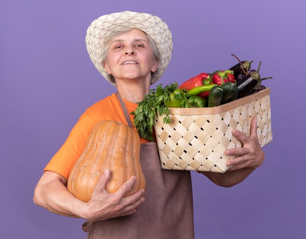 Heureux jardinier femme âgée portant chapeau de jardinage holding potiron et panier de légumes sur violet