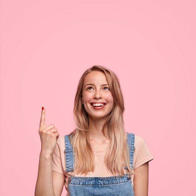 Heureux jeune adolescent femme mignonne en salopette en denim, indique, a un sourire positif, se sent heureux tout en remarquant quelque chose de grand vers le haut