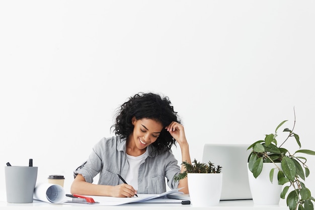 Heureux jeune architecte afro-américain souriant portant chemise grise sur haut blanc faire des dessins