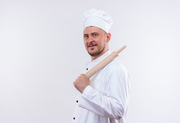 Heureux jeune beau cuisinier en uniforme de chef debout en vue de profil et tenant un rouleau à pâtisserie isolé sur un espace blanc