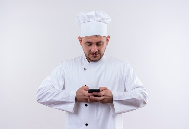 Heureux jeune beau cuisinier en uniforme de chef tenant et utilisant un téléphone mobile isolé sur un espace blanc