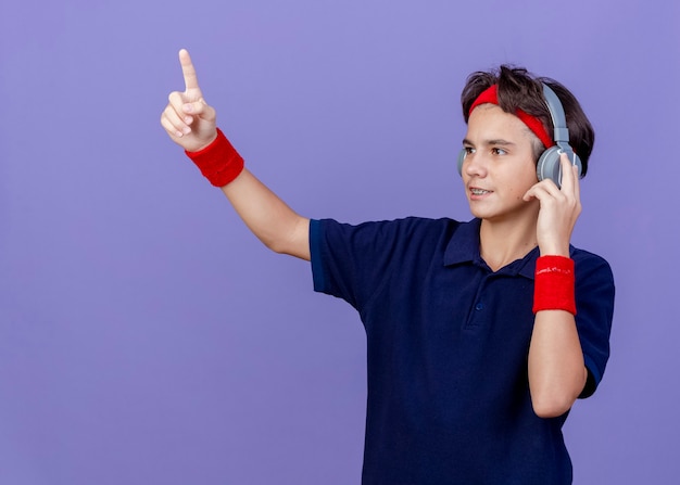 Heureux jeune beau garçon sportif portant un bandeau et des bracelets et des écouteurs avec un appareil dentaire en regardant le côté toucher les écouteurs pointant vers le haut isolé sur fond violet avec espace de copie