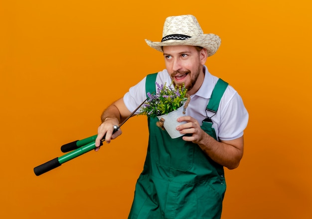 Photo gratuite heureux jeune beau jardinier slave en uniforme et hat holding flowerplant et sécateurs en levant isolé