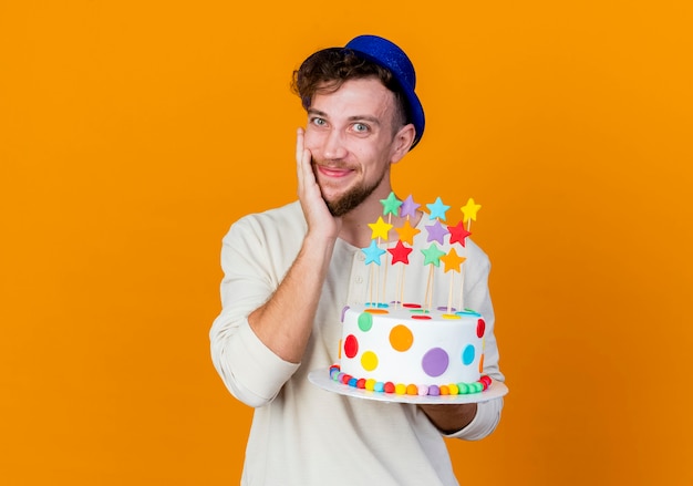 Heureux jeune beau mec de fête slave portant chapeau de fête tenant le gâteau d'anniversaire avec des étoiles regardant la caméra toucher le visage isolé sur fond orange avec espace copie