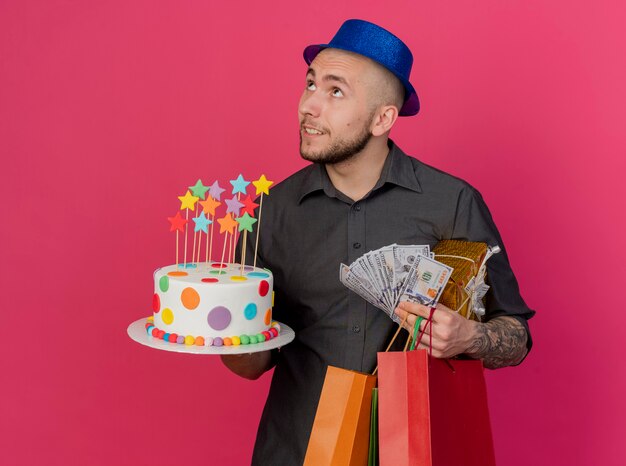 Heureux Jeune Beau Mec De Fête Slave Portant Un Chapeau De Fête Tenant Un Paquet De Cadeau D'argent De Gâteau D'anniversaire Et Des Sacs En Papier Tournant La Tête à L'autre En Levant Isolé Sur Fond Cramoisi Avec Espace De Copie